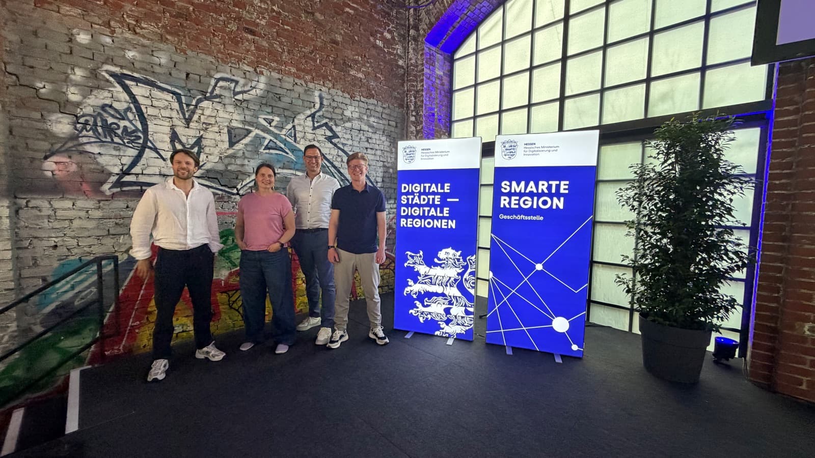 Johannes Reif, Sabrina Bechtle, Hendrik Schaus und Tim Kiesler vor den Bannern des Kongresses „Digitale Städte – Digitale Regionen“ in Marburg